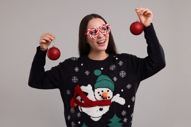 Photo of Happy young woman in Christmas sweater and funny glasses holding festive baubles on grey background
