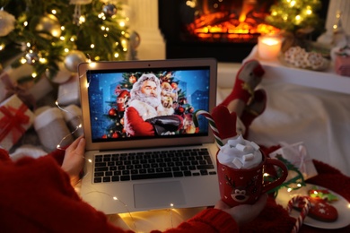 Photo of MYKOLAIV, UKRAINE - DECEMBER 23, 2020: Woman with sweet drink watching The Christmas Chronicles movie on laptop near fireplace at home, closeup. Cozy winter holidays atmosphere
