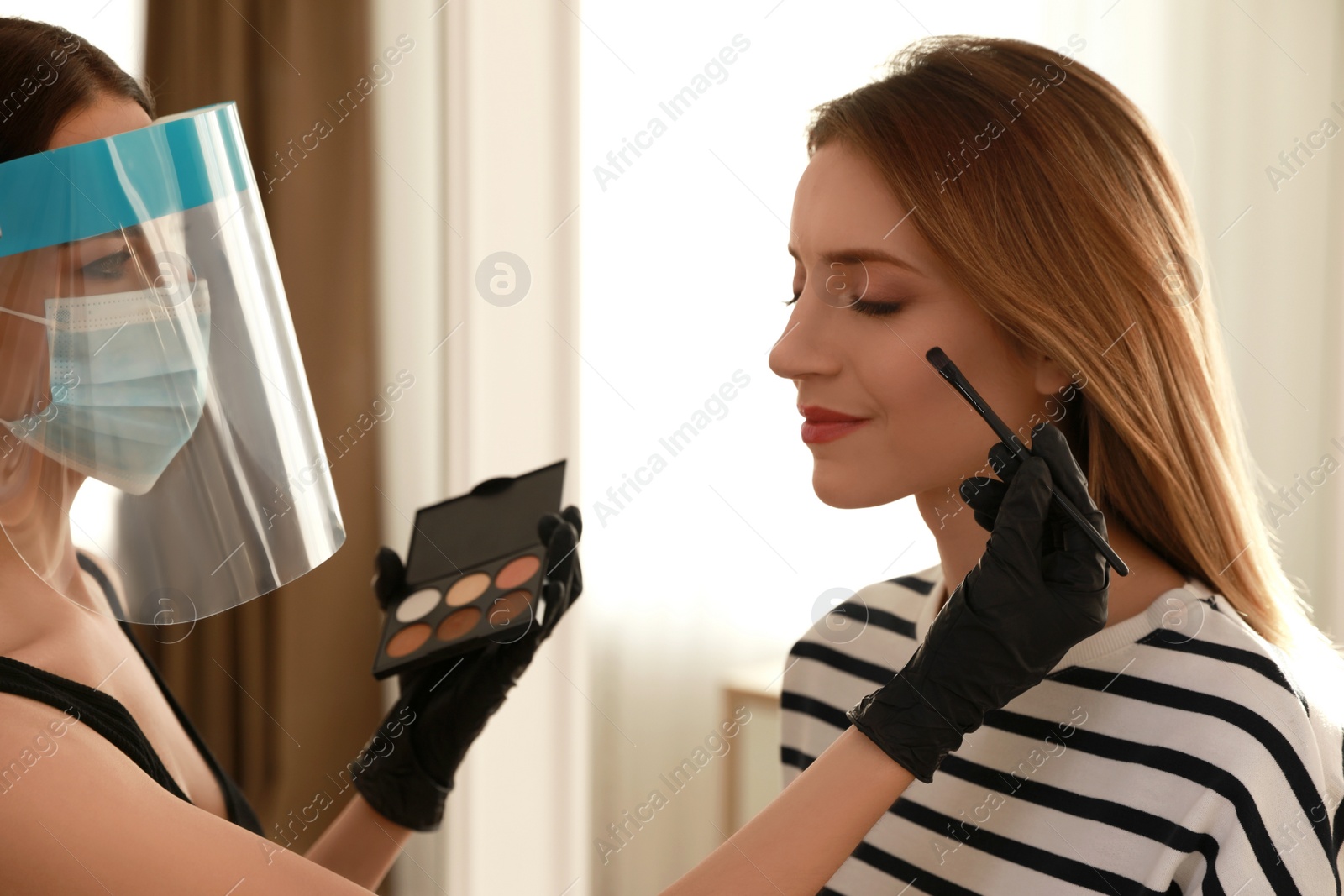 Photo of Makeup artist in protective mask and gloves working with woman indoors. Preventive measures during COVID-19 pandemic