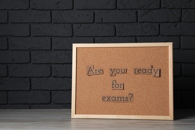 Photo of Cork board with phrase Are You Ready For Exams? on wooden table near black brick wall. Space for text