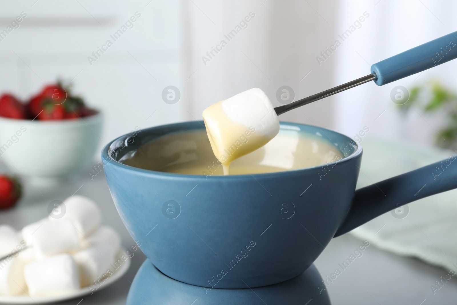 Photo of Dipping marshmallow into pot with white chocolate fondue on table, closeup