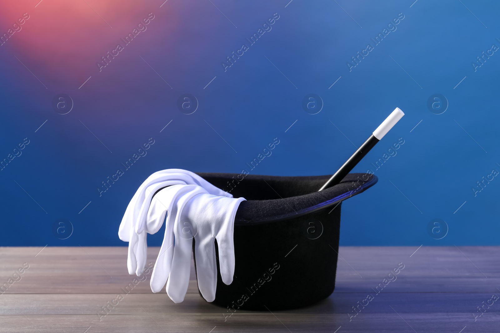 Photo of Magician's hat, wand and gloves on wooden table against color background