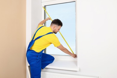 Worker in uniform measuring window with tape indoors. Roller blinds installation