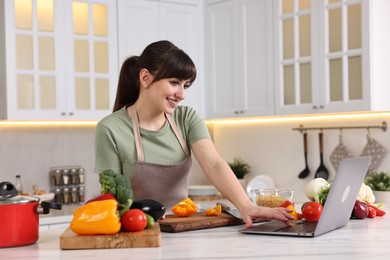 Photo of Happy young housewife using laptop while cooking at white marble table in kitchen