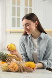 Photo of Woman with lemons and string bag of fresh fruits at light marble table in kitchen