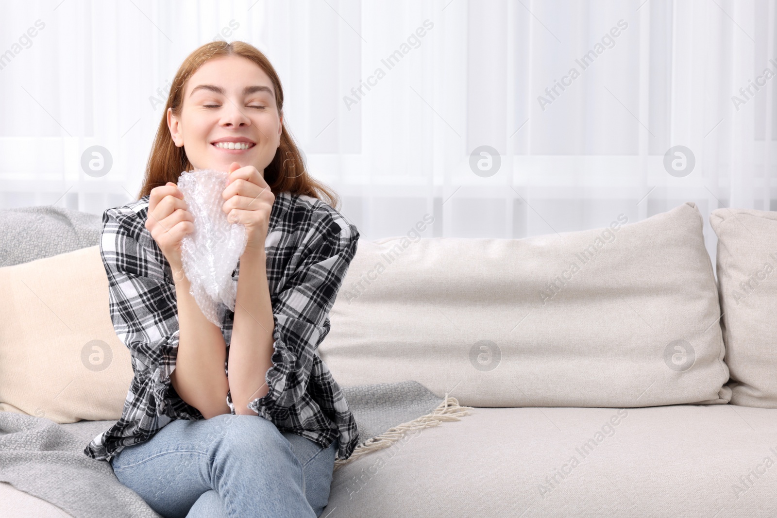 Photo of Woman popping bubble wrap on sofa at home, space for text. Stress relief