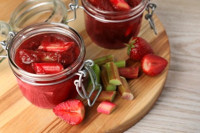 Photo of Jars of tasty rhubarb jam, cut stems and strawberries on white wooden table