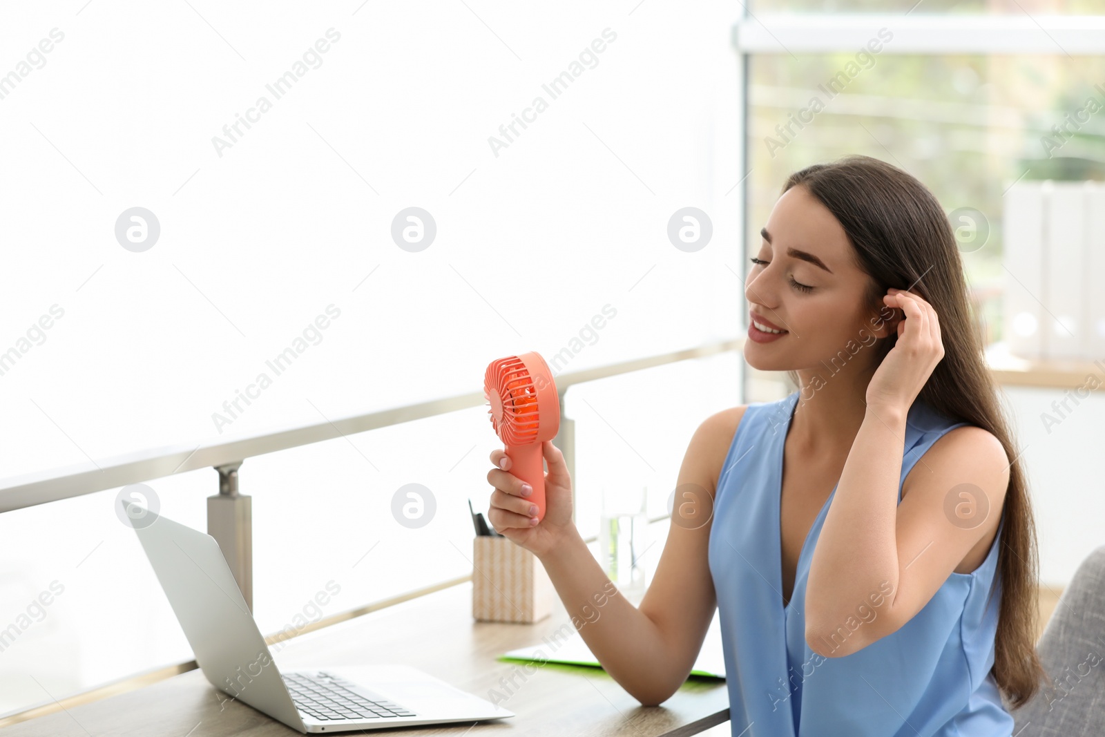 Photo of Young woman enjoying air flow from portable fan at workplace, space for text. Summer heat