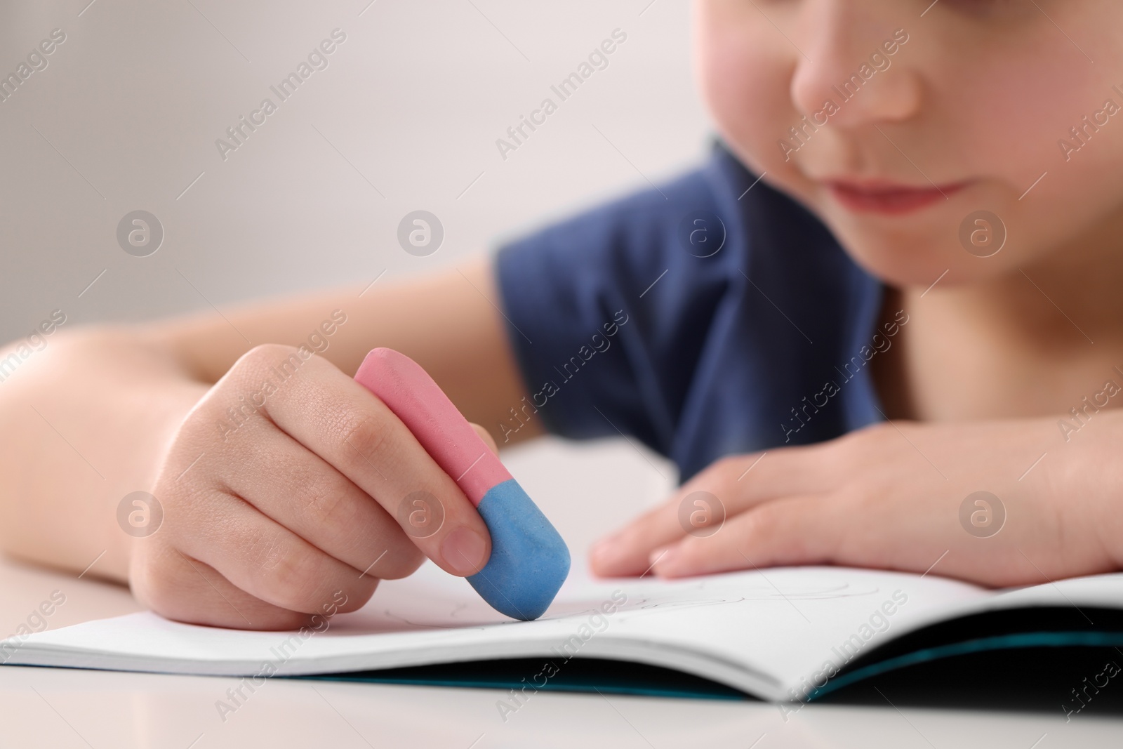 Photo of Little boy erasing mistake in his notebook at white desk, closeup