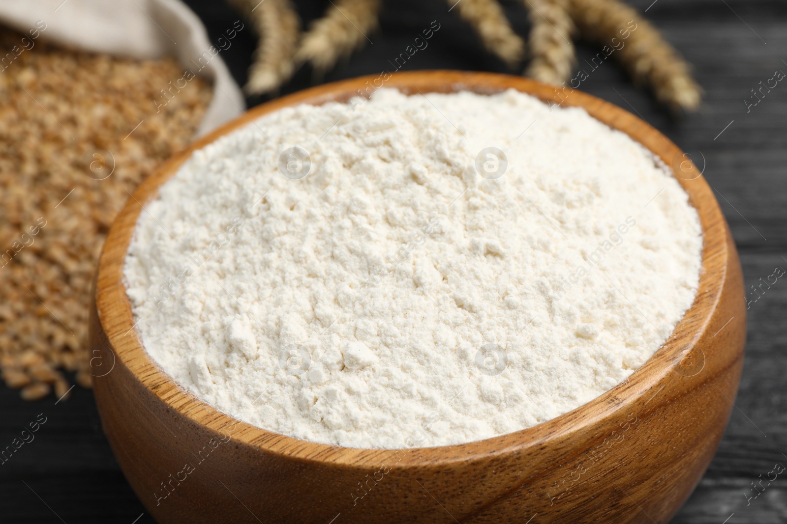 Photo of Wheat flour in wooden bowl on black table, closeup