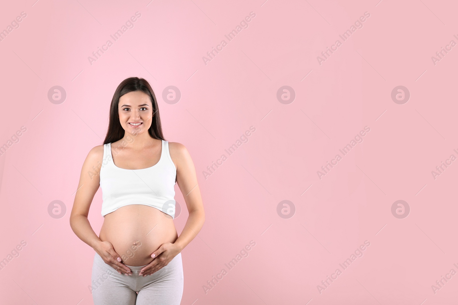 Photo of Beautiful pregnant woman holding hands on belly against color background. Space for text