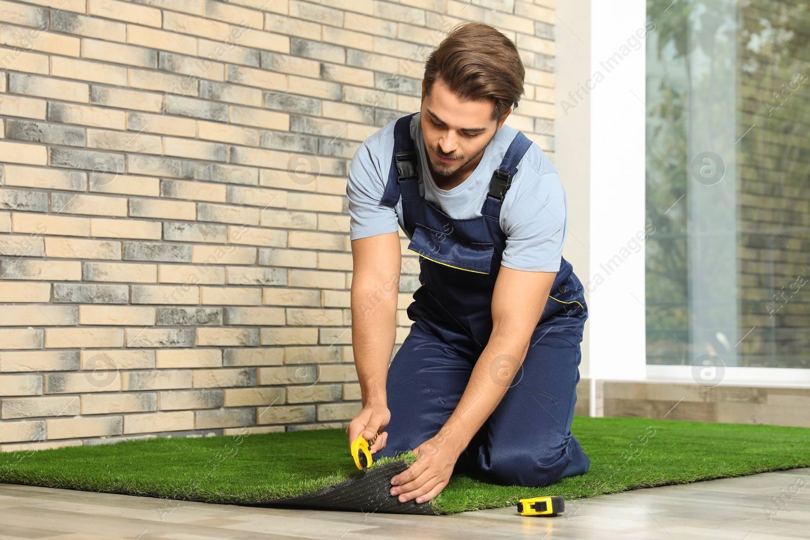 Photo of Man in uniform cutting artificial grass carpet indoors