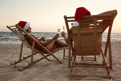 Photo of Lovely couple wearing Santa hats and drinking champagne together on beach. Christmas vacation