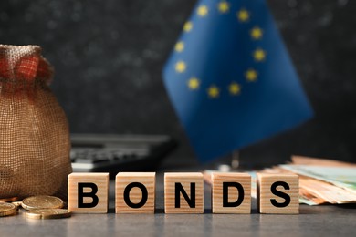 Photo of European Union flag, banknotes and word Bonds made of wooden cubes on grey table