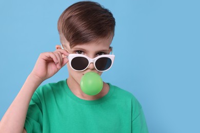 Boy in sunglasses blowing bubble gum on light blue background, space for text