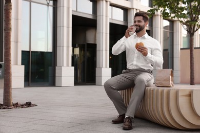 Photo of Businessman with hamburger and paper cup of coffee having lunch on bench outdoors