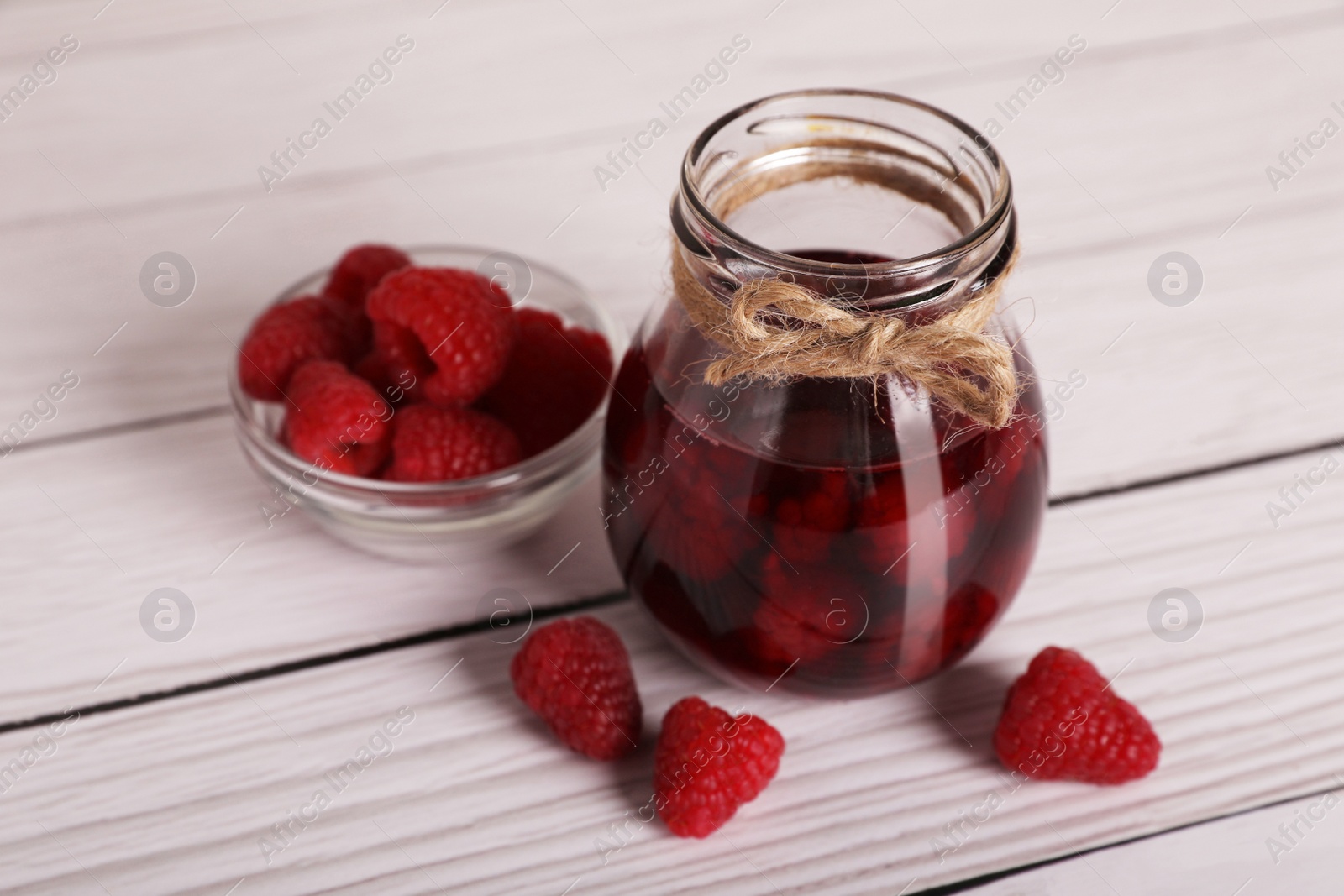 Photo of Jar of tasty canned raspberry jam and fresh berries in glass bowl on white wooden table