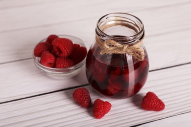 Photo of Jar of tasty canned raspberry jam and fresh berries in glass bowl on white wooden table