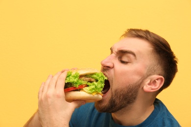 Young man eating tasty burger on color background. Space for text