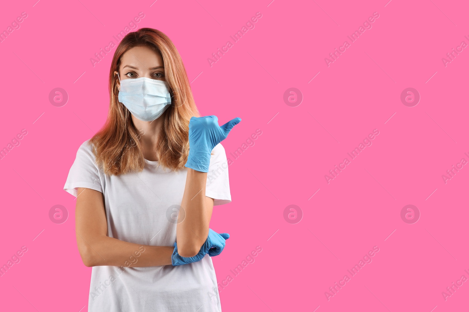 Photo of Young woman in medical gloves and protective mask pointing thumb aside on pink background. Space for text