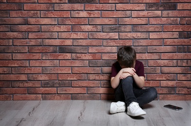 Sad little boy with mobile phone sitting on floor near brick wall. Space for text