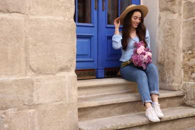 Beautiful woman with bouquet of spring flowers on stairs near building, space for text