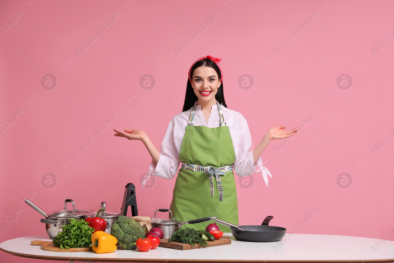 Photo of Young housewife at white table with utensils and products on pink background