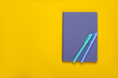 Photo of Closed blue notebook and pens on yellow background, top view. Space for text