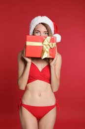 Photo of Sexy young woman in bikini and Santa hat with gift box on red background. Christmas celebration
