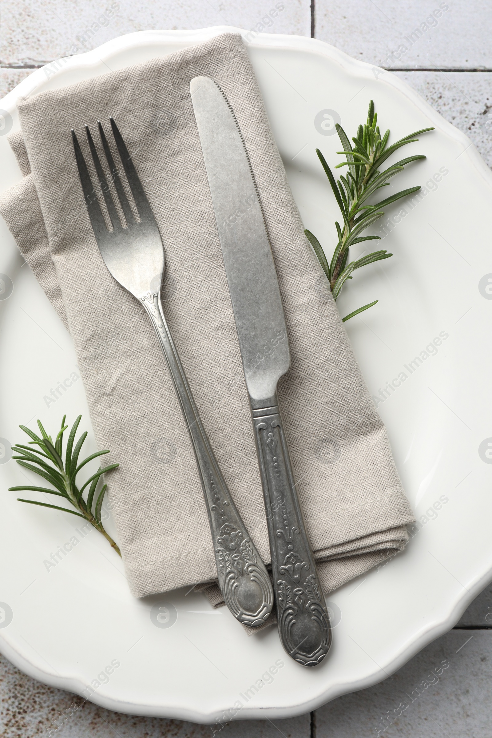 Photo of Stylish setting with cutlery, napkin, rosemary and plate on light tiled table, top view