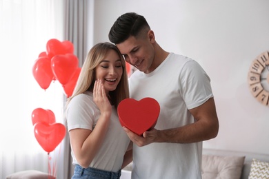 Photo of Man presenting gift to his girlfriend in room decorated with heart shaped balloons. Valentine's day celebration