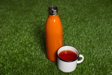 Photo of Modern orange thermos bottle and cup on green grass outdoors