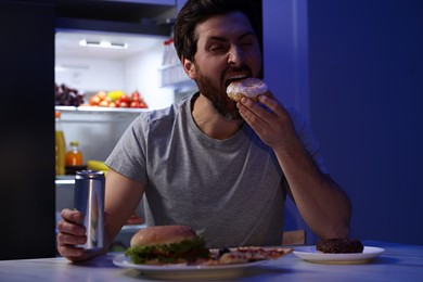 Photo of Man holding tin can and eating donut in kitchen at night. Bad habit