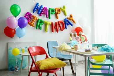 Photo of Table with treats and phrase HAPPY BIRTHDAY made of colorful balloon letters in living room