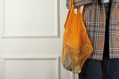 Woman holding mesh bag with different items near white wall, closeup and space for text. Conscious consumption