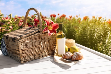 Composition with picnic basket and bottle of wine on white wooden table in lily field. Space for text