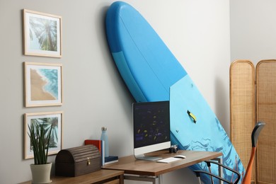 Stylish workplace with modern computer and SUP board near light wall in room. Interior design