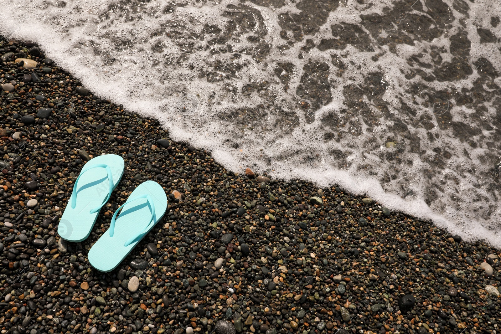 Photo of Stylish turquoise flip lops on pebble beach near beautiful sea wave, above view. Space for text