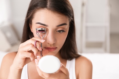 Photo of Attractive young woman curling her eyelashes indoors