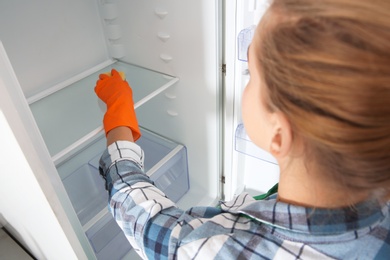 Photo of Woman in rubber gloves cleaning empty refrigerator at home