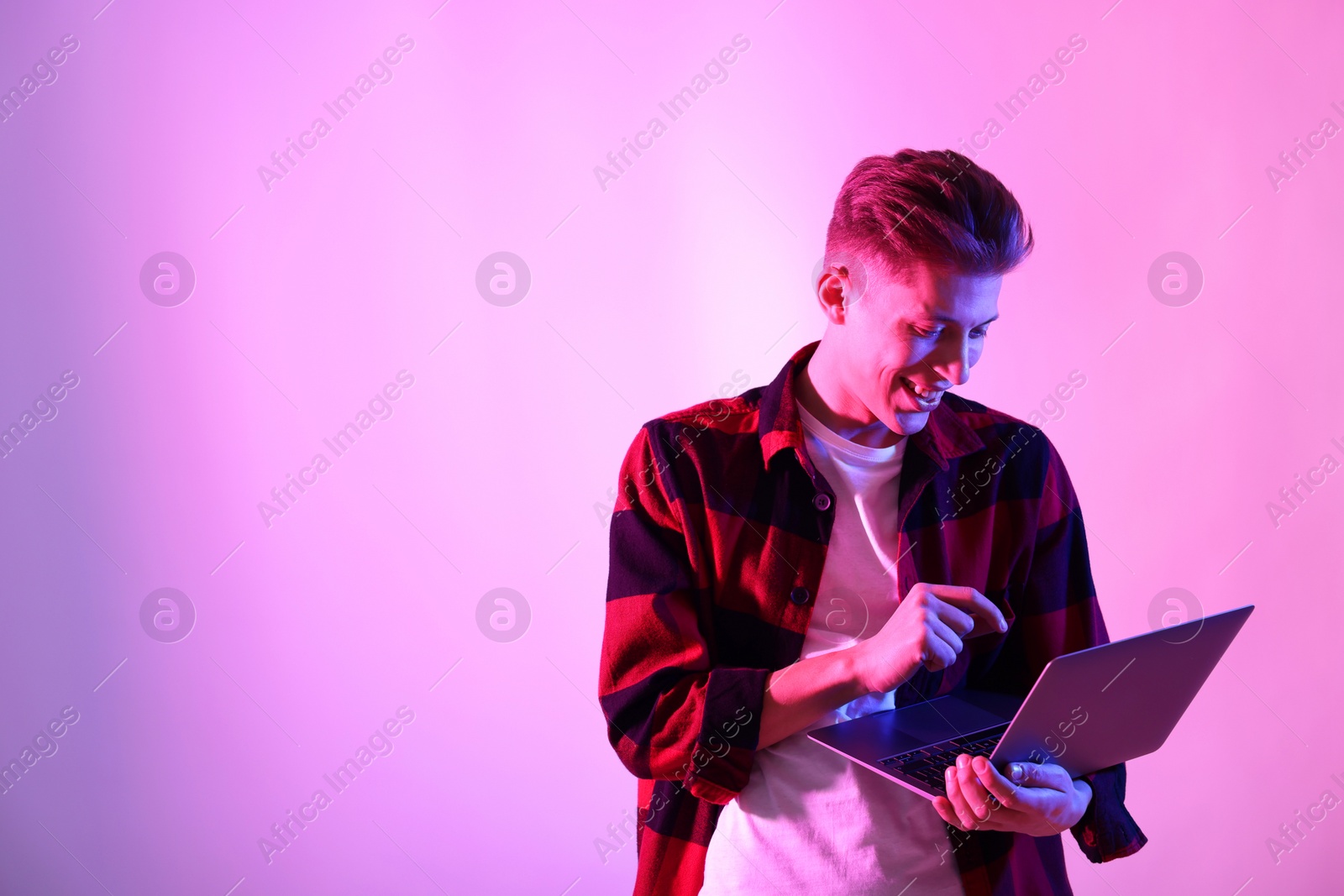 Photo of Young man with laptop talking via video chat on pink background in neon lights. Space for text