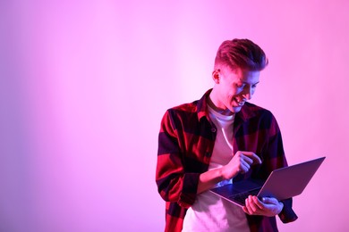 Photo of Young man with laptop talking via video chat on pink background in neon lights. Space for text