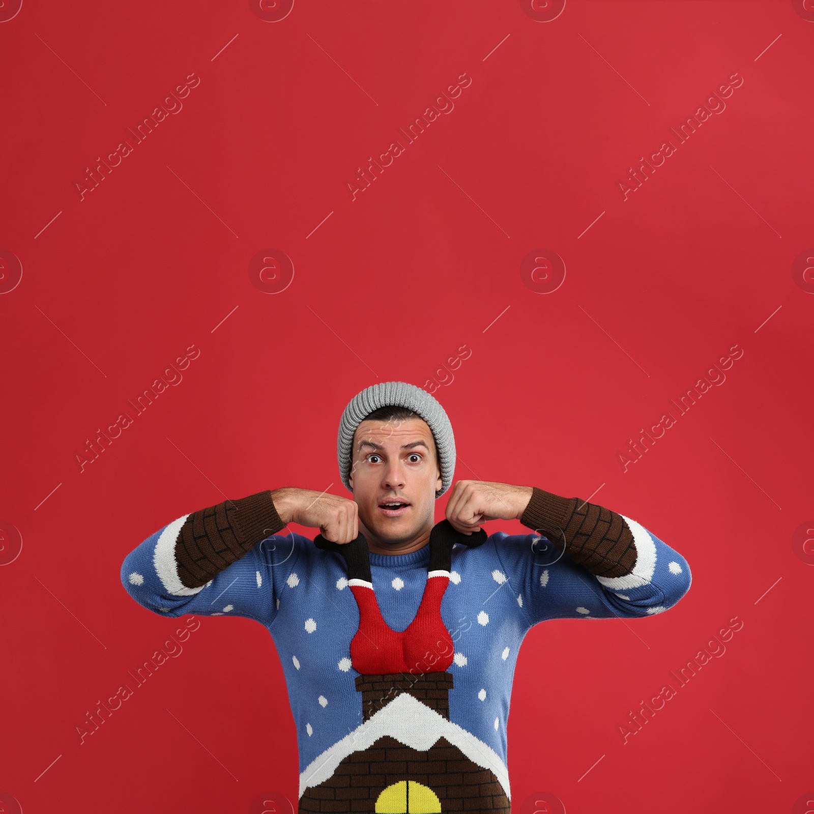 Photo of Surprised man in hat pulling decorative Santa on his Christmas sweater against red background