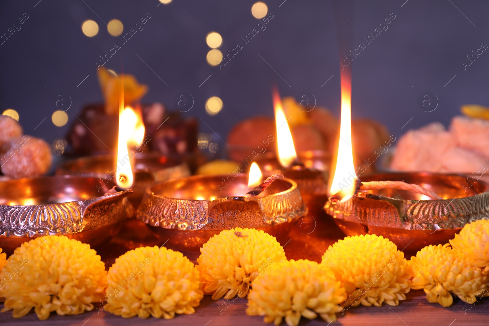 Photo of Diwali celebration. Diya lamps and chrysanthemum flowers on table against blurred lights, closeup