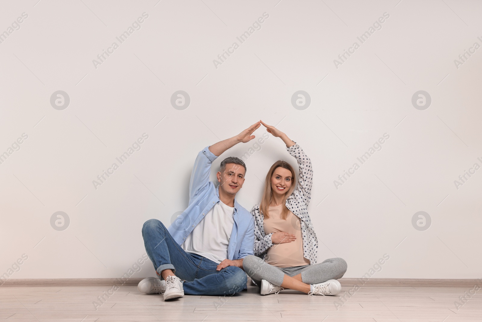 Photo of Young family housing concept. Pregnant woman with her husband forming roof with their hands while sitting on floor indoors