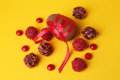 Tasty chocolate egg with red ribbon and candies on yellow background, flat lay