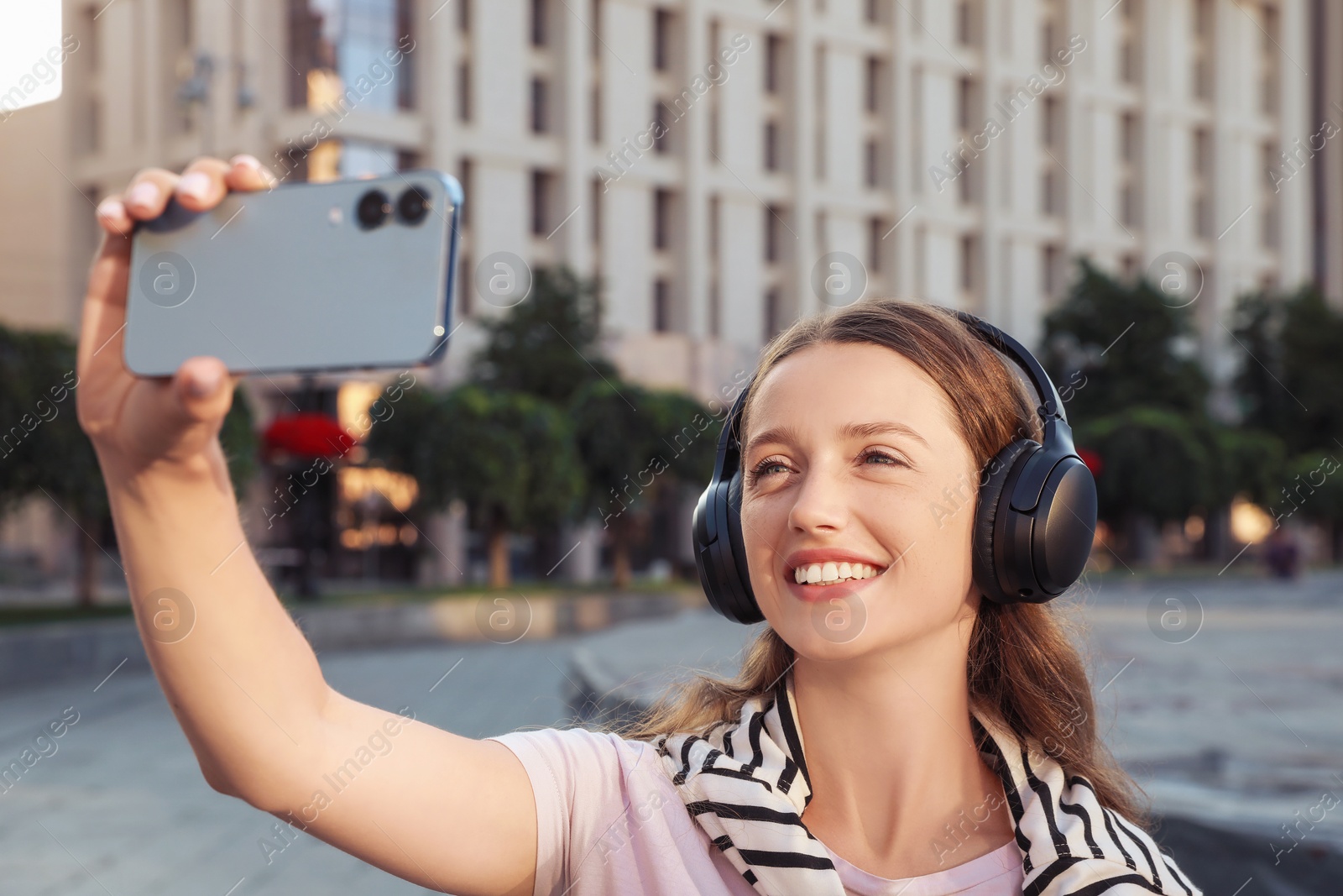 Photo of Smiling woman in headphones taking selfie on city street