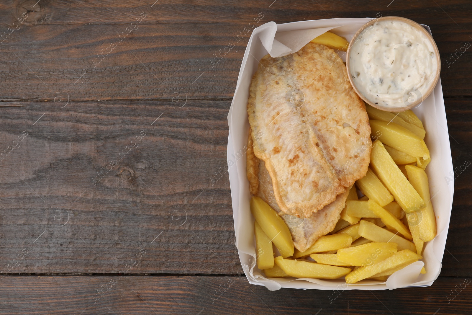 Photo of Delicious fish and chips with tasty sauce in paper box on wooden table, top view. Space for text