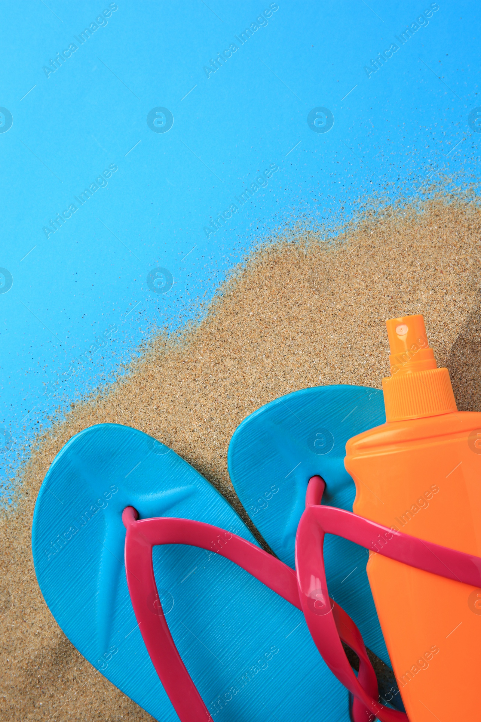 Photo of Flip flops, sunscreen and sand on light blue background, top view with space for text. Beach accessories