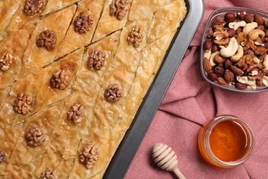 Photo of Delicious baklava with walnuts in baking pan, honey and nuts on red cloth, flat lay